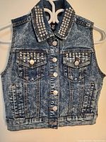 Front view of women's denim vest jacket showing studded collar, chest flap pockets with silver snap buttons, acid wash finish.