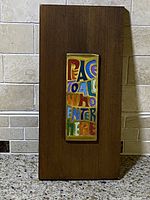 Front view of wooden wall plaque with colorful brass plate reading "PEACE TO ALL WHO ENTER HERE."