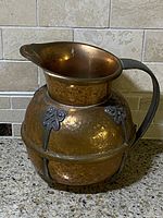 Side view of vintage hammered copper pitcher with dark metal handle and floral decorative motifs