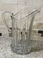 Clear vintage crystal pitcher with vertical ribbed design and starburst base photographed against tiled backsplash, showing curved handle and pouring spout from front angle.
