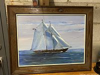 Framed original painting showing two-masted sailing ship on the ocean against a cloudy sky. Wooden frame with gold inner trim.