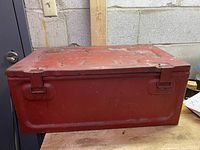 Top view of red rectangular metal military box with two latches on front, showing paint wear and vintage condition.