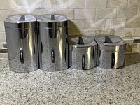 Four Kromex stainless steel canisters lined up on a kitchen counter. Two larger canisters for flour and sugar and two smaller canisters for coffee and tea each with matching lids featuring black handles. Labels on canisters read Flour, Sugar, Coffee, and Tea.