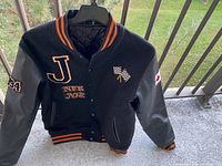 Full front view of a black vintage varsity jacket with leather sleeves and various patches including a letter 'J', racing flags, and the number '94'. The jacket has striped ribbed collar, cuffs, and hem.