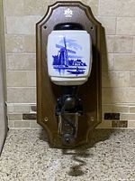 Front view of the wall-mounted coffee grinder showing the ceramic container with blue windmill design and wooden backing plaque