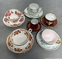 Top view of multiple vintage English teacups and matching saucers displaying patterns and colors including pink roses, green garlands, deep red with gold, orange floral, and blue floral designs.