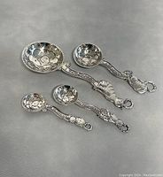 Full view of the four metal measuring spoons with cat-shaped handles and embossed fish designs in bowls, washed and displayed on grey surface.