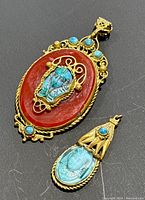 Close view of two brass and turquoise pendants with carved face motifs, showing red-orange base on larger pendant.