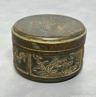 Closed bronze round box showing hand-etched floral and calligraphy decorations on the sides and top.