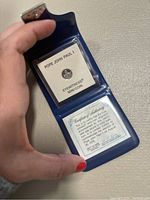 Photo showing the open blue wallet containing the coin and certificate of authenticity with fingers holding it.