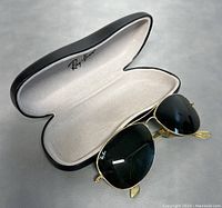 Open Ray-Ban black hard case with white lining holding gold frame aviator sunglasses with dark green lenses beside it.