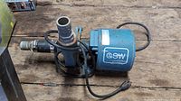 Side view of blue GSW electric water pump lying on wooden surface, showing motor housing, pipes, hoses, and power cord.