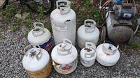 Photo of 7 propane tanks of different sizes, mostly white with rust spots, arranged outdoors on gravel.