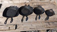 Photo of four cast iron pans on a wooden surface including one square grill pan and three round frying pans of different sizes