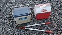 Photo shows two small portable coolers and a tree pruner placed on gravel. The coolers are from Coleman and Little Playmate brands, and the pruner has red handles and a metal cutting section.