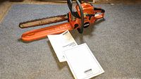 Chainsaw laid flat with bar scabbard and two manual booklets on gray carpet