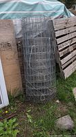 Roll of welded wire fencing leaning against wooden panels outside on the ground in a grassy area.