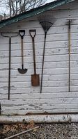 Photo of 5 garden hand tools hanging against white wooden siding: garden cultivator, hoe, spade, leaf rake, mattock with wooden handles and metal heads.