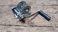 Top view of the metal hand winch showing crank handle with black grip and gear mechanism on wooden surface.