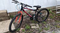 Full side view of the kids mountain bike showing black frame with Mongoose branding, red front forks, knobby tires, handlebars with grips, black saddle, back tire flat and detached from ground, placed on stone and soil surface.