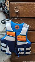 Photo of a blue, white, and orange Body Glove kids life jacket vest hanging on a hanger. Close up shows zipper front, two quick-release buckles, and brand logo along chest.