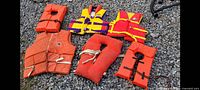 Overview of six older life jackets arranged on gravel. Includes multiple colors and styles, showing signs of use.