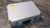 Closed Plano silver aluminum case with black handle and yellow sticker on lid, placed on wood surface.