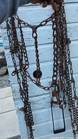 Photo showing a rusty metal tire chain held up near a white wooden wall, highlighting the chain links and hooks.