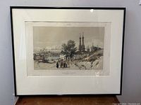Front view of framed lithograph print showing village scene with church spires and figures. Includes visible title and artist text on print.
