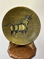 Front view of a round decorative ceramic wall plate depicting an abstract bull on a textured glazed background, placed on a wooden display stand.
