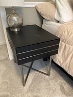 Photo showing black night stand placed beside a bed with beige bedding and a lamp on top, highlighting the drawer and metal legs.