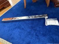 Silver rectangular metallic silver wall shelf on blue carpeting, alongside sealed mounting hardware packet.