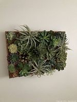 Rectangular decorative wall piece densely covered with various faux succulents in green and slight purple tones, arranged in a garden-style layout.