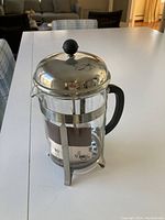 Glass French press coffee maker on table showing metal frame, clear glass carafe, black handle, and silver lid with knob.