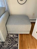 Light blue upholstered hassock ottoman positioned next to a matching sofa with wooden floor and patterned rug visible.
