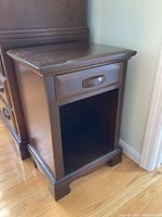 Solid wood nightstand with one drawer, open lower shelf, dark brown finish, on hardwood floor against light-colored wall.