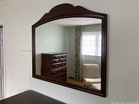 Photo shows large rectangular mirror with dark wood frame mounted on a wall, reflecting part of a bedroom including a chest of drawers, chair, window, and curtains.