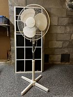 White standing oscillating fan with four-legged base, placed in front of a window frame against a gray brick wall.