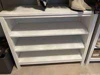Photo showing one white painted wooden low bookcase with three shelves, placed on carpeted floor with shoes and other items on top and nearby