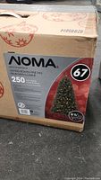 Box of Noma Lighted Bowen Pine Tree with product details showing 250 mini lights, 6.5 foot height, and inclusion of replacement bulbs and tree stand.