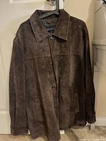 Front view of a brown suede men's jacket with button closure, collar, and pockets, hanging on a hanger against a white door.