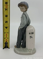 Side view of porcelain figurine showing boy in blue clothing leaning against white milestone with measuring tape indicating height.