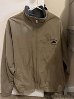 Beige XL jacket hanging on hanger, front view showing zipper and embroidered logo.