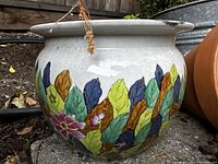 Side view of the white ceramic flower pot showing colorful hand-painted leaves and a pink flower design around its body. Wide rim and bulbous round shape.