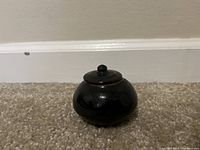 Front view of a small, glossy black ceramic jar with a lid and round compact shape on carpeted floor.