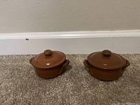 Image showing two small brown ceramic pots with lids placed on carpet against a beige wall. Both pots are similar in design with side handles and round knobs on lids.