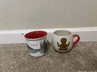 Two holiday themed ceramic mugs placed on carpet against a white wall. One mug has black pine tree silhouettes and 'cozy up' text with red interior, the other has a gingerbread man design with a red handle.