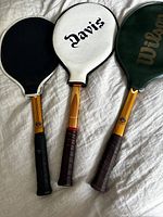 Three vintage wooden tennis rackets with covers – black cover for one Imperial racket, white cover labeled 'Davis' for Dunlop racket, and green Wilson cover for the other Imperial racket