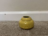 Front and side view of a small yellow ceramic vase with textured pattern and glossy finish on carpeted floor against wall.