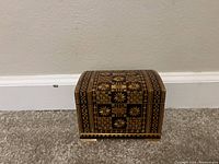 Small wooden treasure box with detailed geometric and floral marquetry patterns on lid and sides, shown against a white baseboard and carpeted floor.